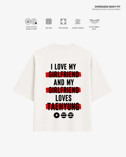 An oversized white boxy-fit T-shirt shown from the back, featuring the text 'I LOVE MY GIRLFRIEND AND MY GIRLFRIEND LOVES TAEHYUNG' in black with red horizontal highlight bars. Small fabric feature icons and text appear above the shirt on a light gray background.