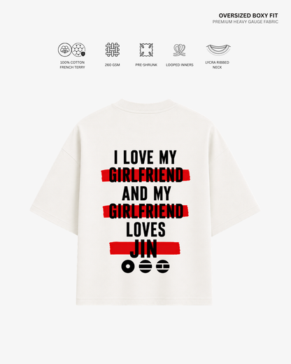 A white oversized boxy-fit T-shirt shown from the back, featuring a bold graphic print in black and red text that reads 'I LOVE MY GIRLFRIEND AND MY GIRLFRIEND LOVES JIN' with red scribble-style bars over key words. Small fabric feature icons and text appear above the shirt on a light gray background.