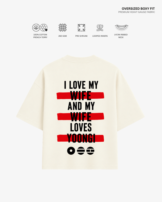 An oversized cream-colored short-sleeve t-shirt with a bold graphic on the back reading 'I LOVE MY WIFE AND MY WIFE LOVES YOONGI' in black text over red horizontal stripes, shown on a light gray background with fabric feature icons and text at the top.